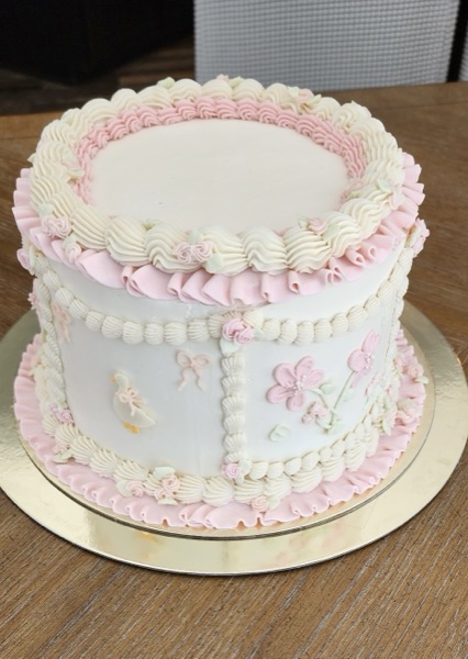Elevated three-quarter view of a Lambeth-style baby shower cake in white and pink with a painted yellow duckling on the front panel, piped pink florals on the side panel, ruffled borders, and pink mini-rose accents, on a gold board
