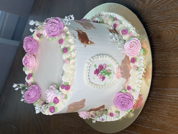 Single-tier white cake with a crown of pink buttercream roses, a piped rose-bouquet cameo medallion on the side, copper palette-knife strokes, and a pink rose border at the base, on a gold board