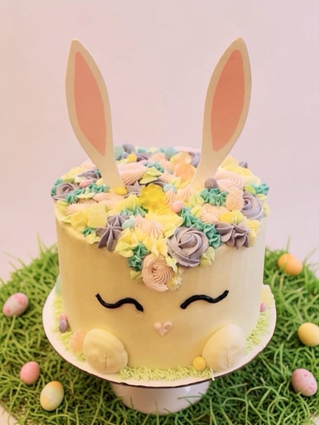 Pastel Easter bunny cake — sleepy buttercream face with paper ears, candy egg feet, and a pastel buttercream floral crown on top