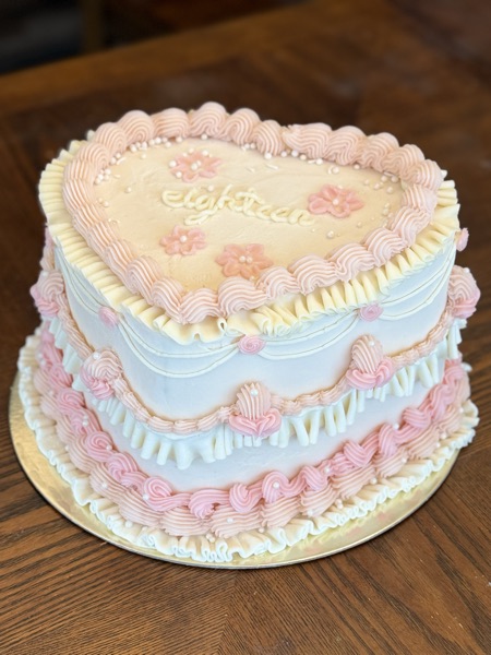 Elevated view of a heart-shaped Lambeth cake in soft pink and cream with "eighteen" piped in script on top, surrounded by pink flowers, ruffled borders, and pearl accents