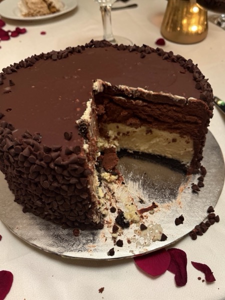 Tall layered chocolate cake with a cheesecake middle, ganache top, and mini chocolate chips coating the sides, with a wedge cut to show the layers