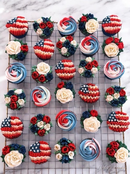 A cooling rack of red, white, and blue patriotic buttercream cupcakes — piped American flags with star florals, tricolor rose swirls, and floral bouquets of cream roses, red mini-roses, and navy blooms, made for the July 4th, 2026 semiquincentennial
