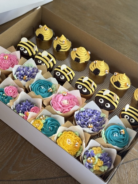 A box of assorted cupcakes: half piped as buttercream bumblebees with yellow and black stripes and fondant wings, the other half as floral bouquets in pink rose, teal and purple hydrangea, and yellow rose wrapped in kraft paper