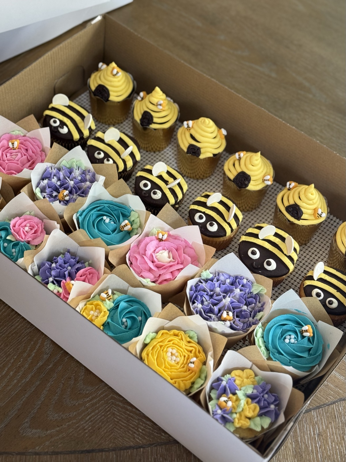 A box of assorted cupcakes: half piped as buttercream bumblebees with yellow and black stripes and fondant wings, the other half as floral bouquets in pink rose, teal and purple hydrangea, and yellow rose wrapped in kraft paper