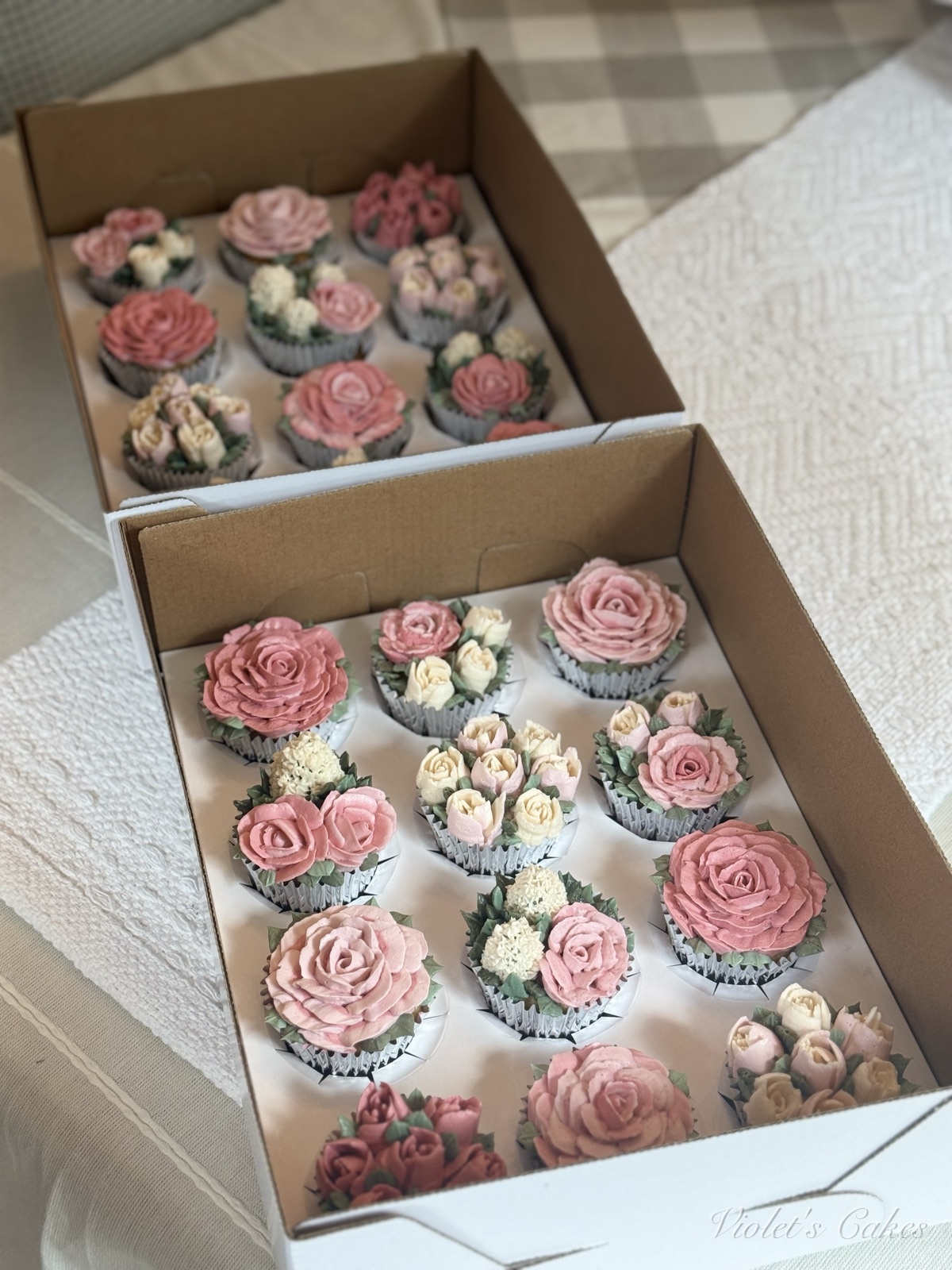 Two boxes of blush and pink buttercream flower cupcakes stacked at an angle — piped roses, tulips, and cream hydrangea florals