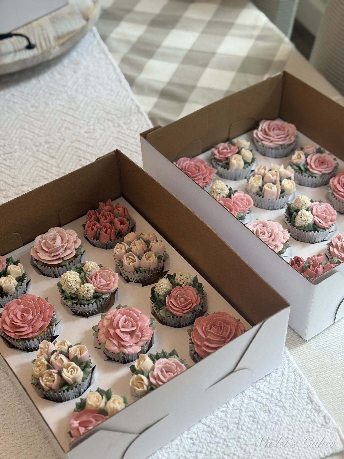 Two open boxes of blush and pink buttercream flower cupcakes on a checkered table — a mix of piped roses, tulips, and cream hydrangea blossoms