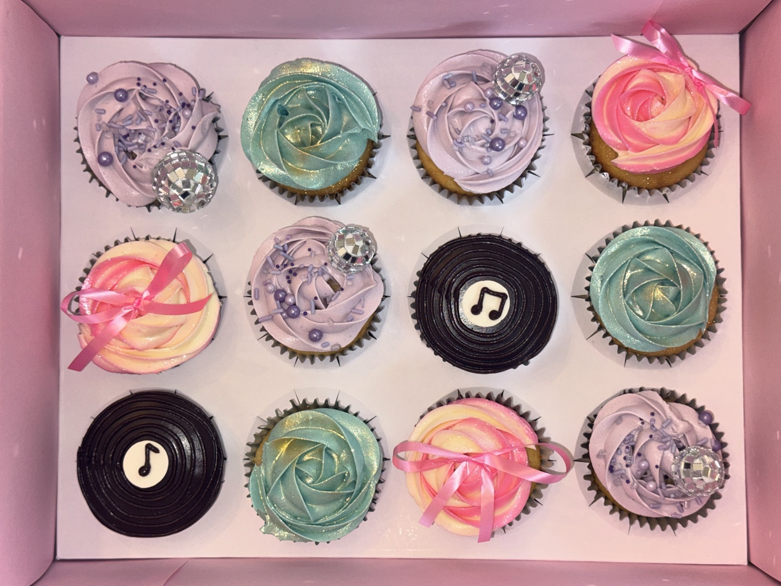 Top-down view of a pink cupcake box with twelve music-themed cupcakes: chocolate vinyl-record cupcakes with white music-note centers, pastel rosette swirls in mint, lavender and pink, with mini disco-ball toppers and ribbon-bow accents