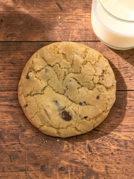 Large soft chocolate-chip cookie on a wooden surface next to a glass of milk