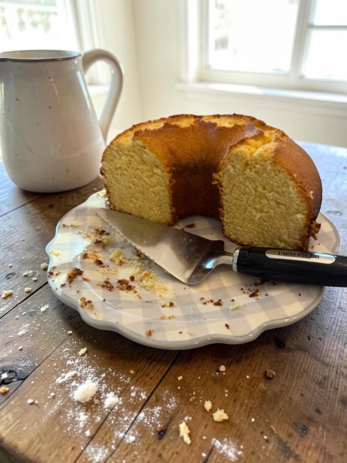 Golden bundt coffee cake with a slice removed, on a gingham scalloped plate with a cake server and pitcher on a wooden table