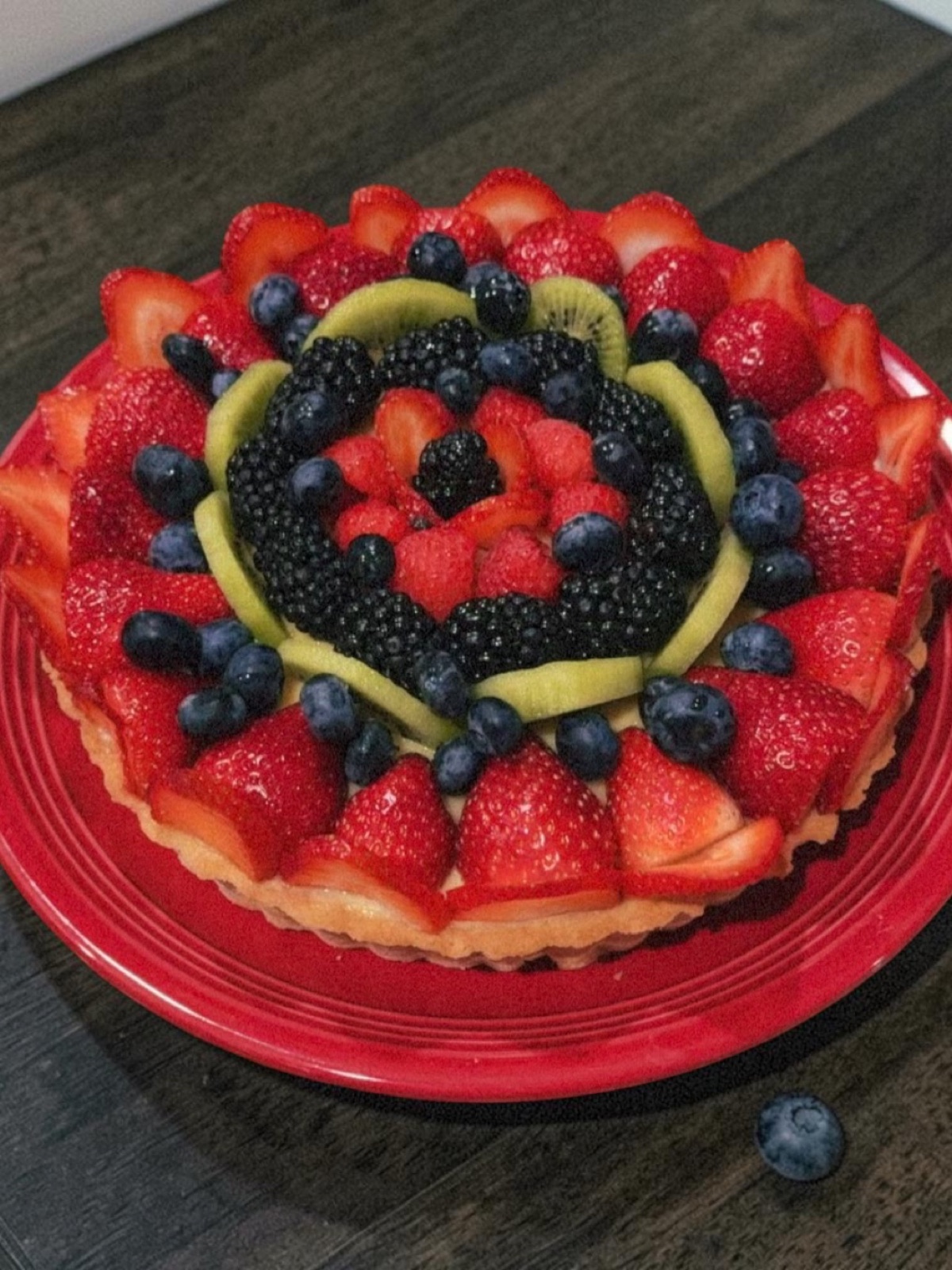 Fruit tart with strawberry, blackberry, blueberry, and kiwi arranged in concentric rings on a red plate