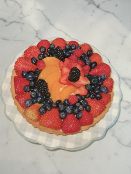 Fruit tart on a glass pedestal with strawberries radiating outward in a sunburst, a ring of blueberries and blackberries, and a vanilla custard center