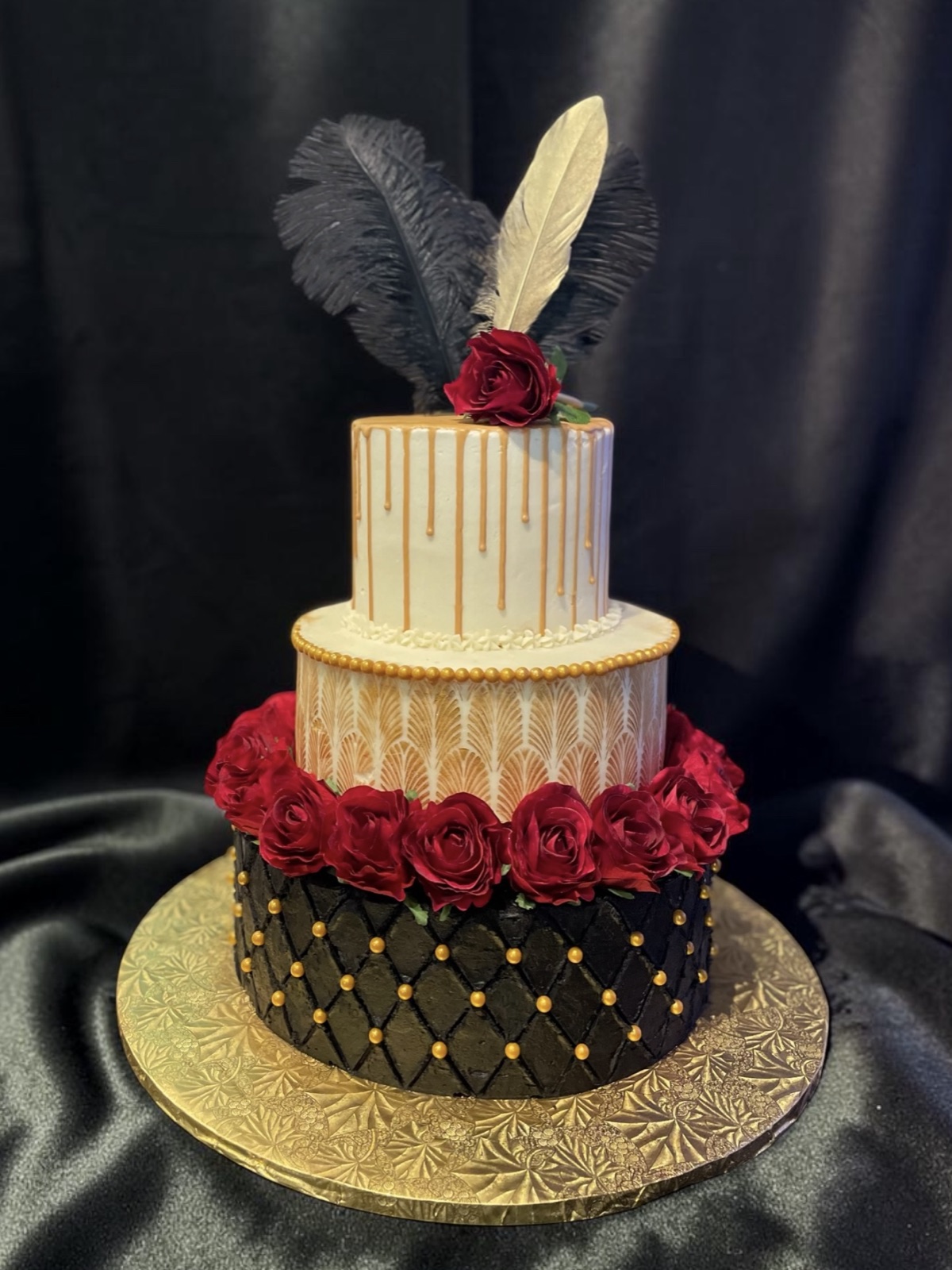 Three-tier art deco cake with a gold-dotted black quilted base, ivory pearled middle tier, ivory drip top with gold drips, a ring of deep red roses, and black and ivory feathers as a topper