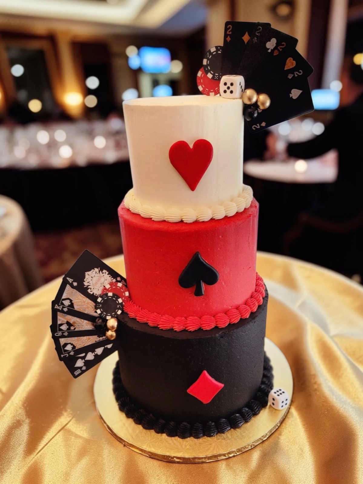 Three-tier ivory, red, and black playing-card cake with heart, spade, and diamond suits, card fans, dice, and a poker chip, on a gold tablecloth