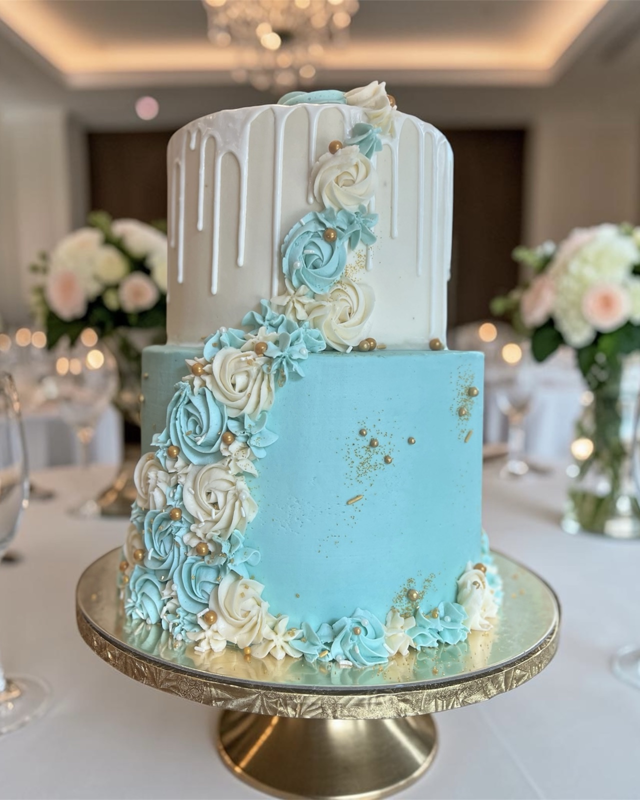 Two-tier teal and ivory cake with white drip top, cascading buttercream roses, and gold pearls, on a gold cake stand at a reception venue
