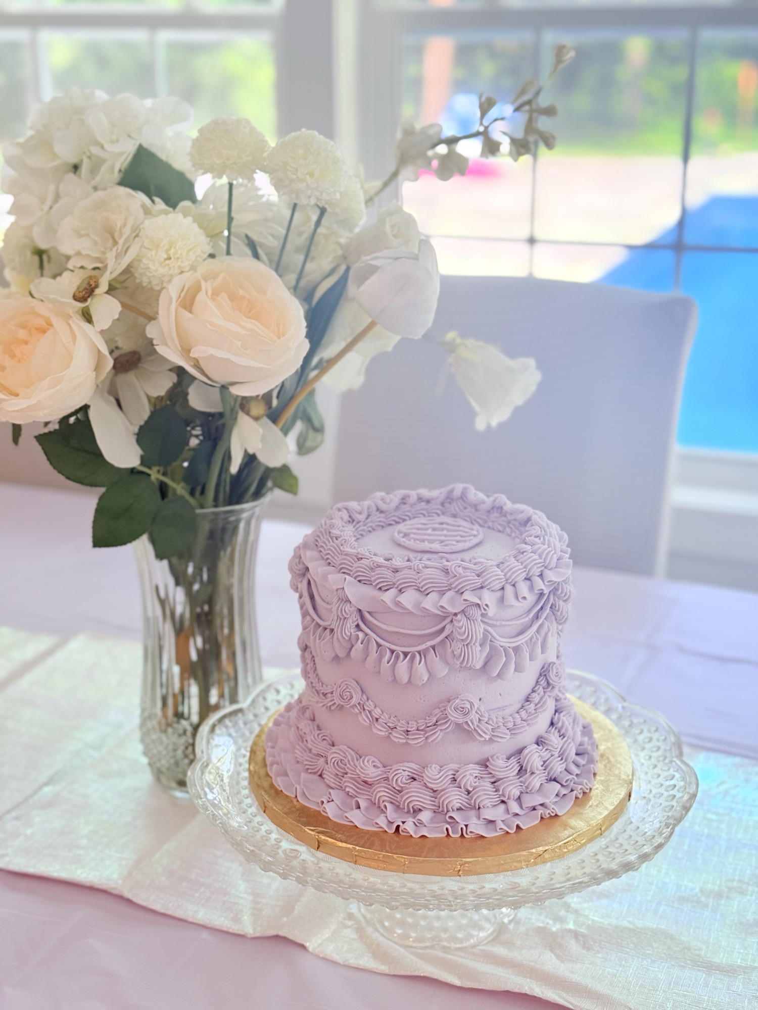 Tall lavender vintage cake on a glass cake stand beside cream roses